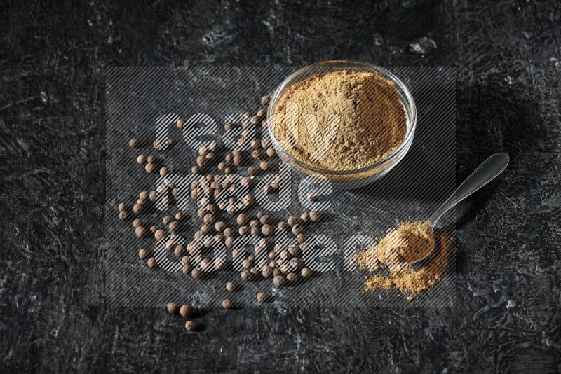 A glass bowl and metal spoon full of allspice powder and whole balls spreaded on a black flooring