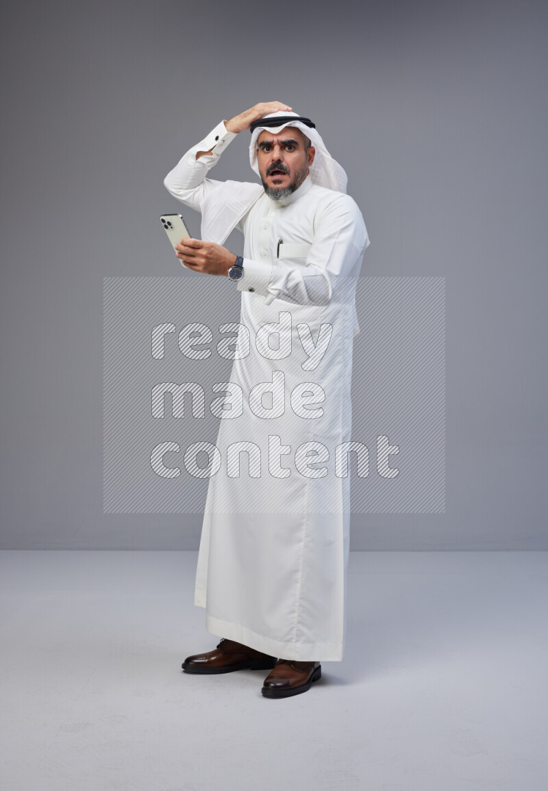 Saudi man Wearing Thob and white Shomag standing texting on phone on Gray background