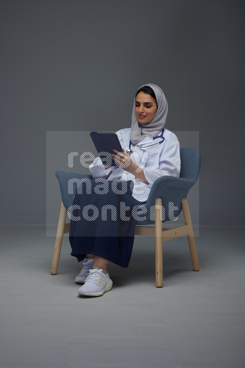 A female doctor wearing a light gray head scarf sitting on a dark grey chair holding an electronic device and pointing to different directions eye level on a grey background
