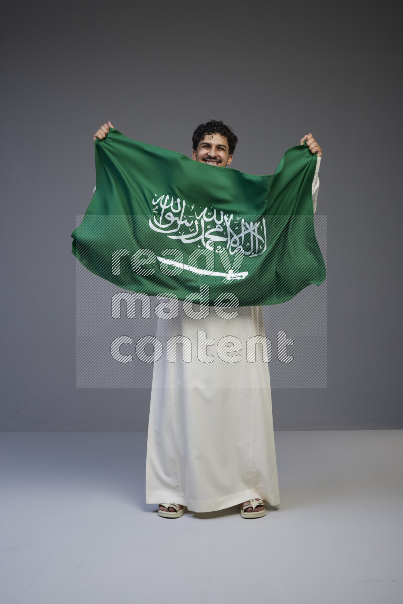 A Saudi man standing wearing thob holding big Saudi flag on gray background