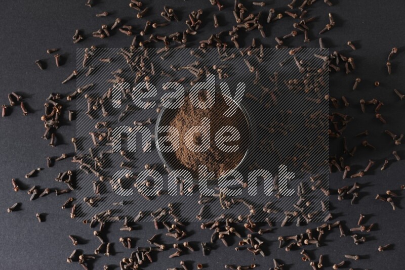 A black pottery bowl full of cloves powder and cloves whole grains on a black flooring