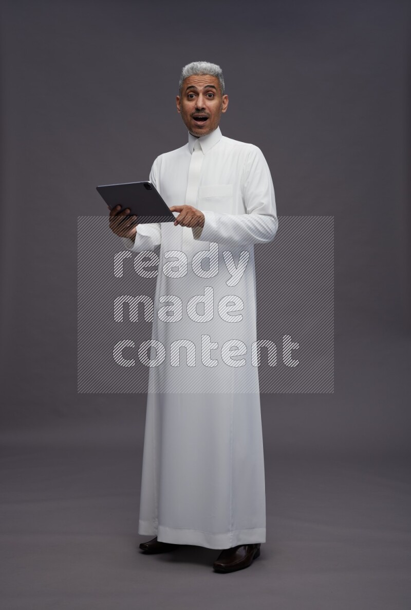 Saudi man wearing thob standing working on tablet on gray background