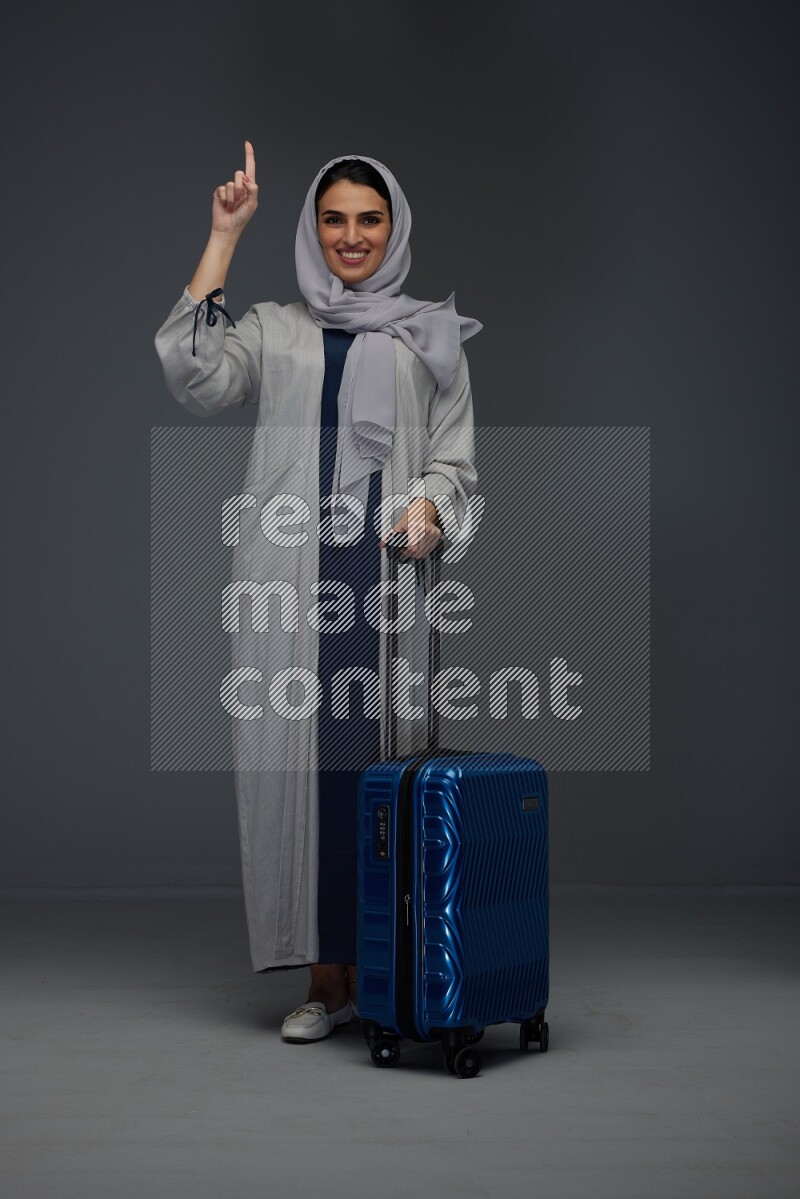 A Saudi woman wearing a light gray Abaya and head scarf standing while holding blue luggage eye level on a grey background