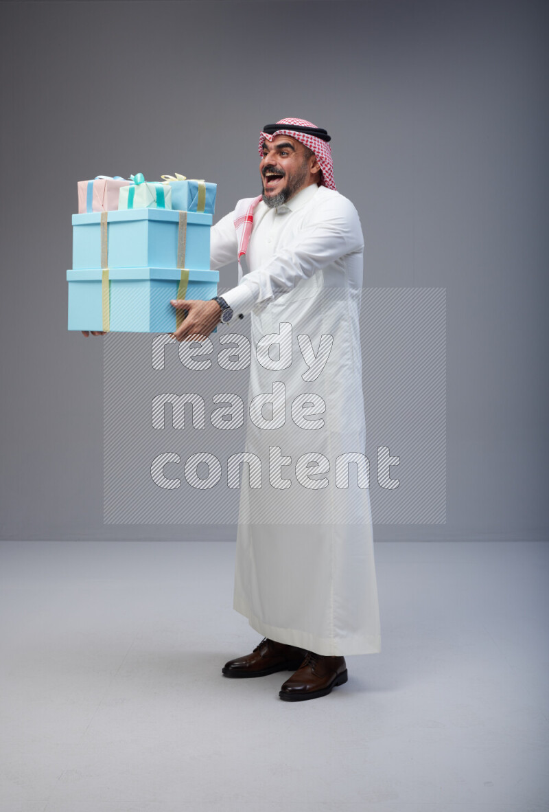 Saudi man Wearing Thob and red Shomag standing holding gift box on Gray background