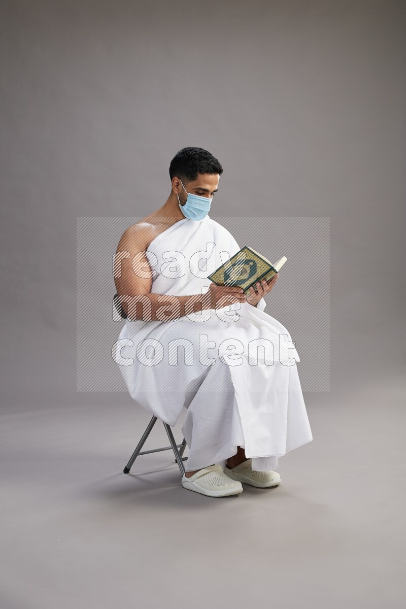 A man wearing Ehram with face mask sitting on chair reading quran on gray background