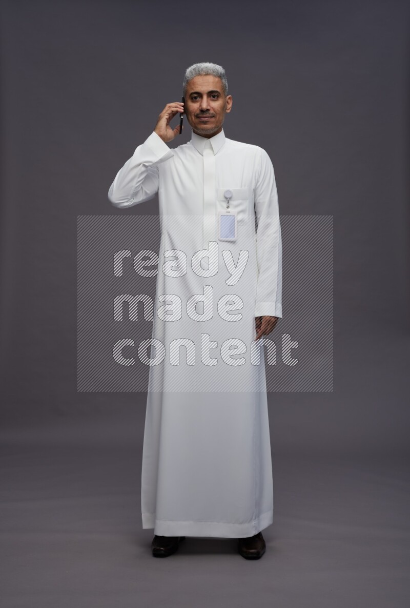 Saudi man wearing thob with pocket employee badge standing talking on phone on gray background
