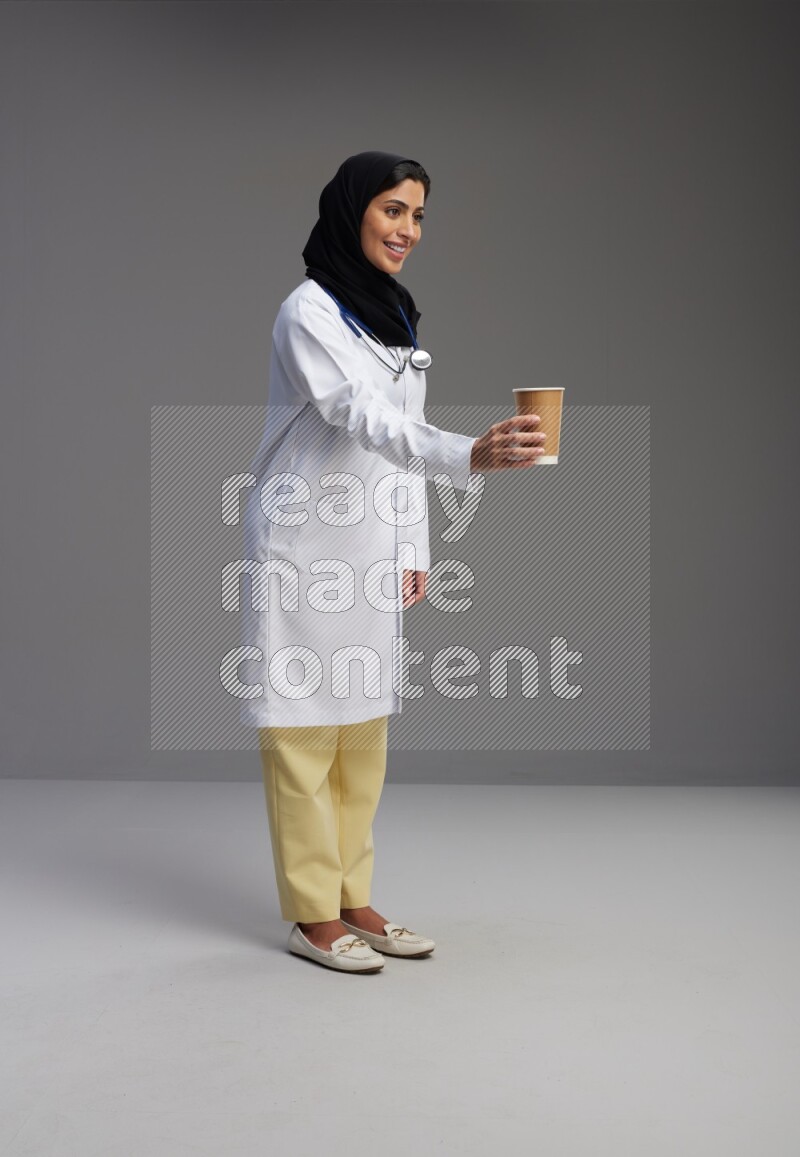Saudi woman wearing lab coat with stethoscope standing holding paper cup on Gray background