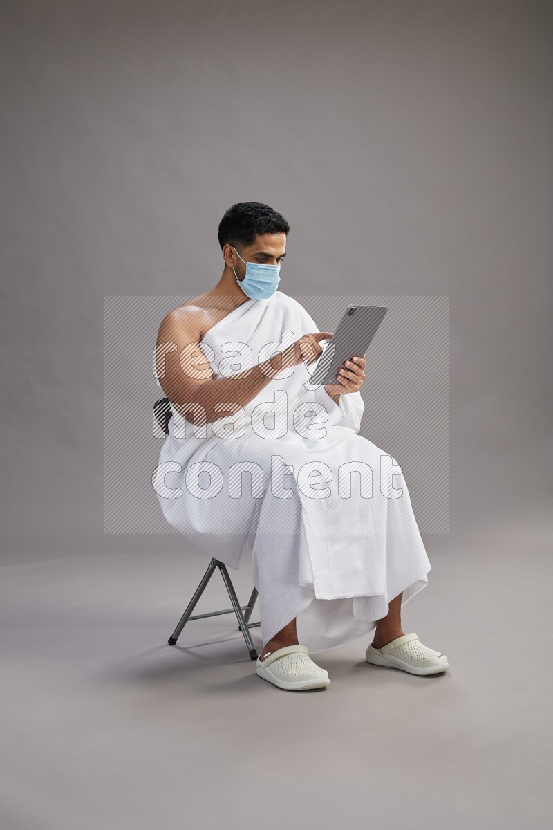 A man wearing Ehram with face mask sitting on chair holding a tablet on gray background