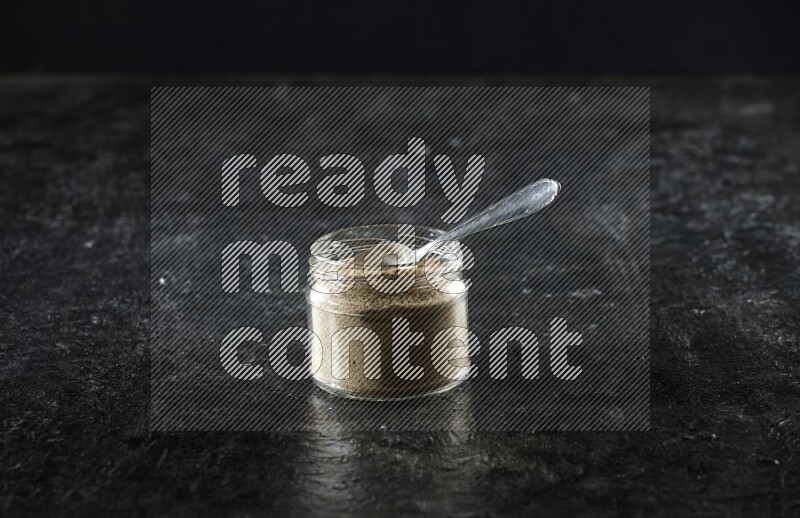 A glass jar and metal spoon full of cardamom powder on textured black flooring