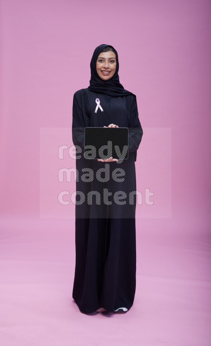 Saudi woman wearing pink ribbon on Abaya standing showing a tablet to camera on pink background