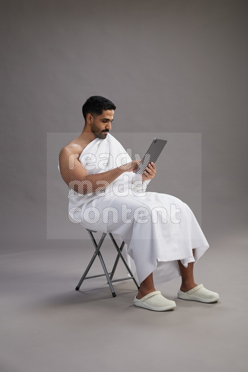 A man wearing Ehram sitting on chair holding a tablet on gray background