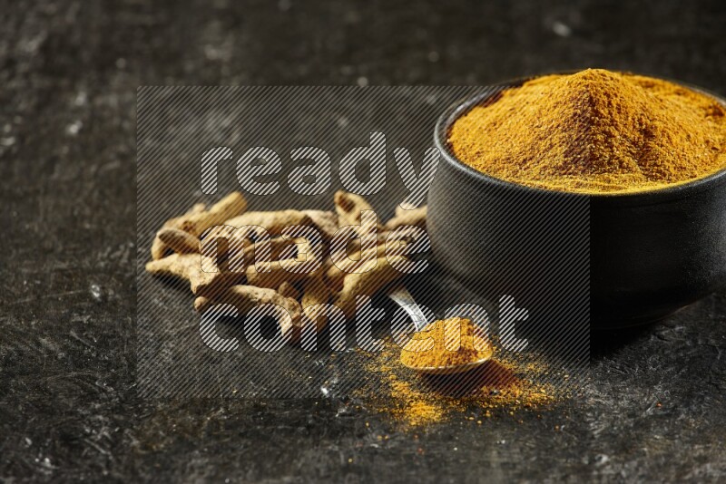 A pottery black bowl and a metal spoon full of turmeric powder with dried turmeric fingers on a textured black flooring