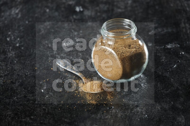 A glass spice jar and a metal spoon full of cumin powder on a textured black flooring