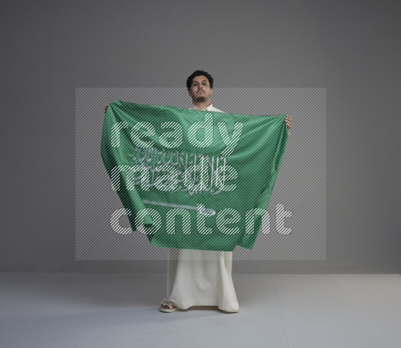 A Saudi man standing wearing thob holding big Saudi flag on gray background