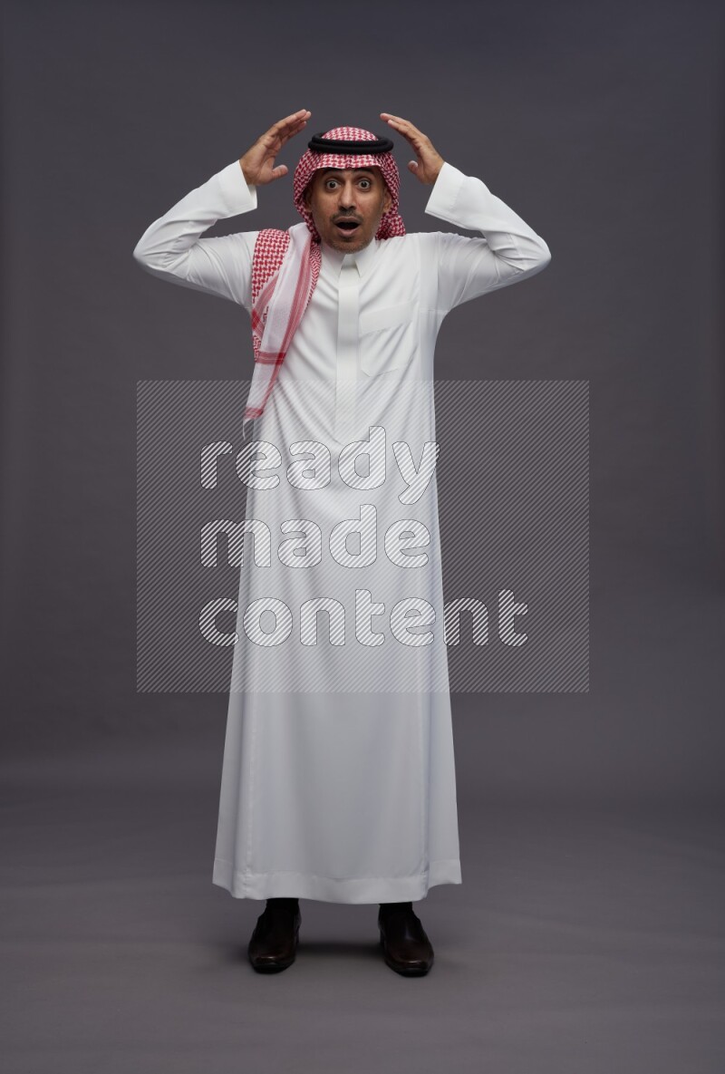 Saudi man wearing thob and shomag standing hands behind head on gray background