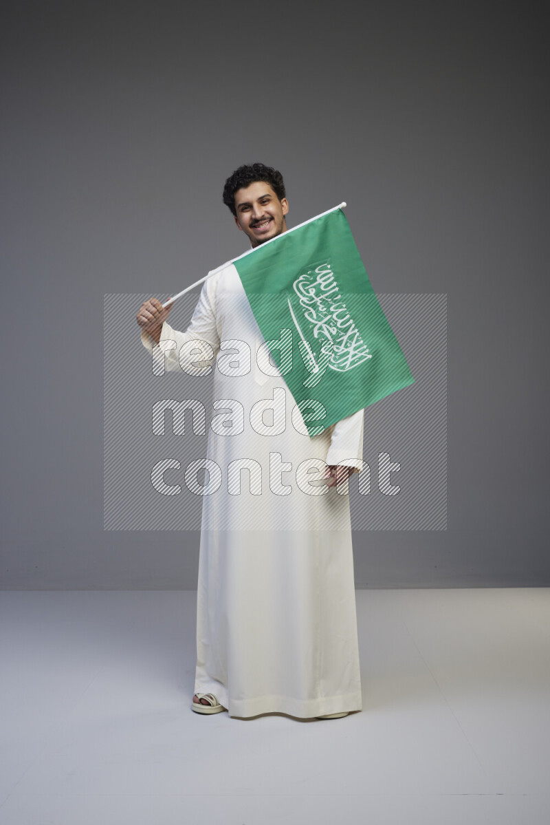 A Saudi man standing wearing thob holding small Saudi flag on gray background