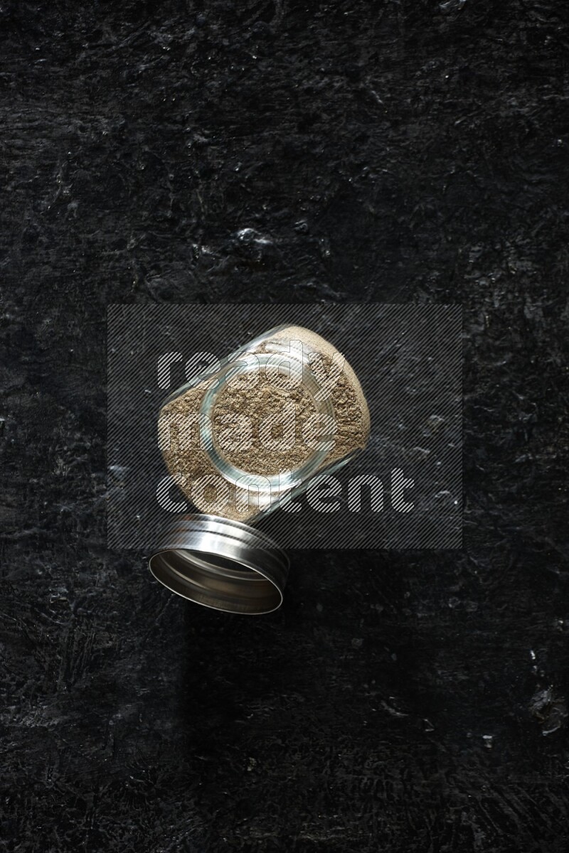 A glass spice jar full of cardamom powder on textured black flooring
