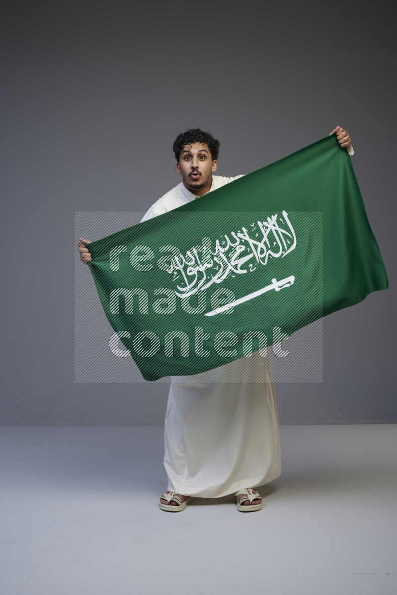 A Saudi man standing wearing thob holding big Saudi flag on gray background