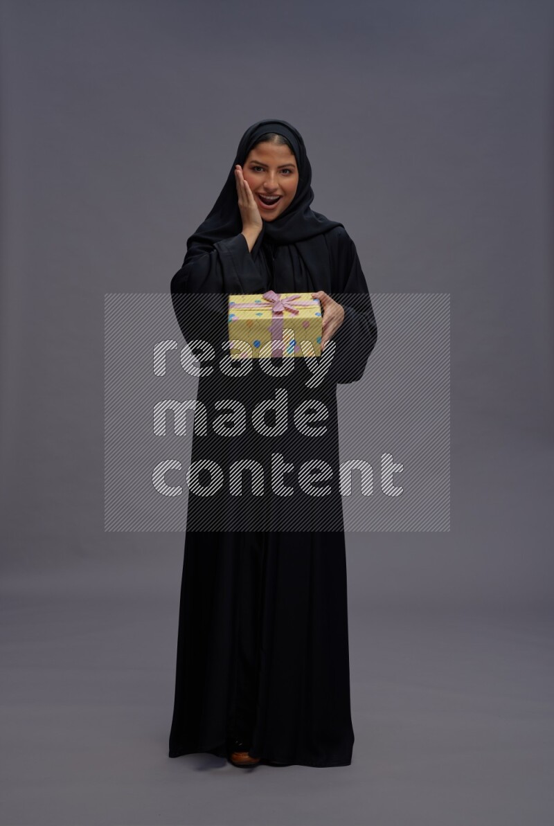 Saudi woman wearing Abaya standing holding gift box on gray background