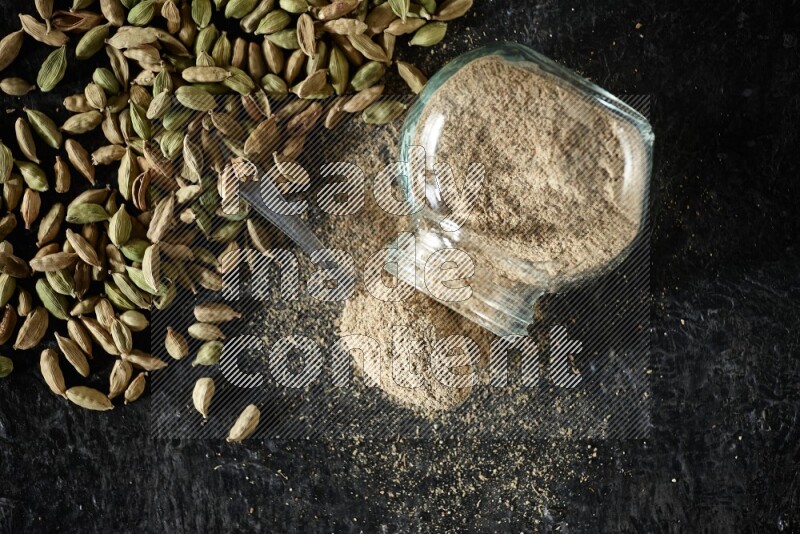 A flipped glass spice jar and a metal spoon full of cardamom powder and cardamom seeds spreaded on textured black flooring