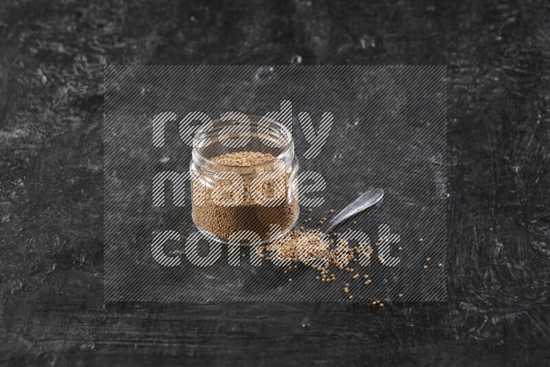 A glass jar and a metal spoon full of mustard seeds on a textured black flooring