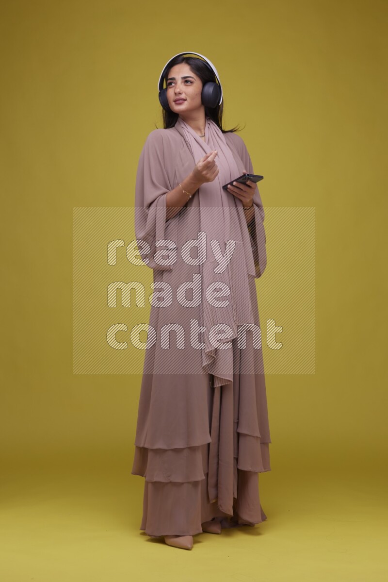 A Saudi woman Listing to Music on a Yellow Background wearing Brown Abaya