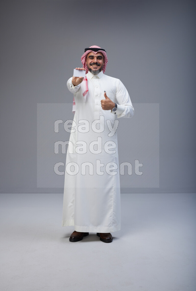 Saudi man Wearing Thob and red Shomag standing holding ATM card on Gray background