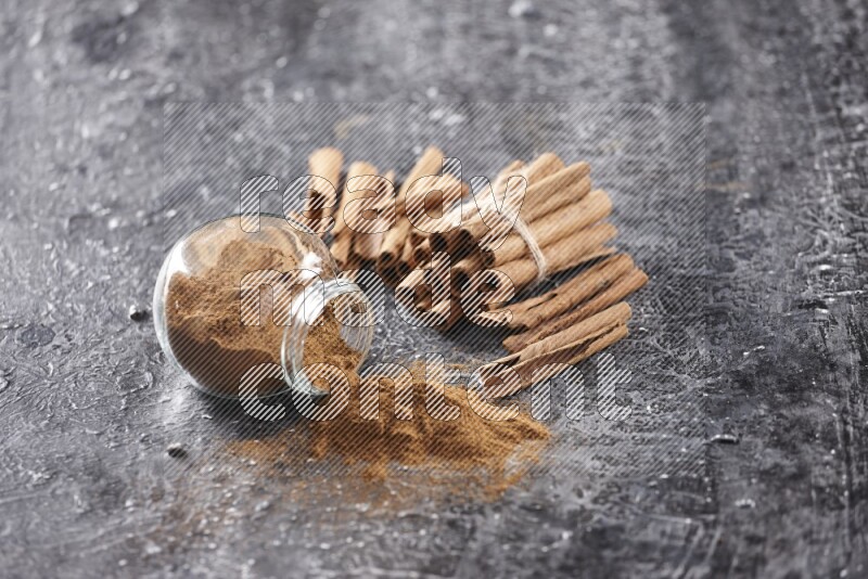 Herbal glass jar full of cinnamon powder flipped with cinnamon sticks stacked and bounded on a textured black background