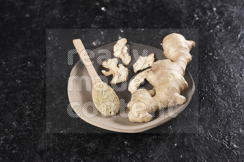 A wooden spoon full of ground ginger powder with fresh and dried ginger, all on a pottery plate on black background