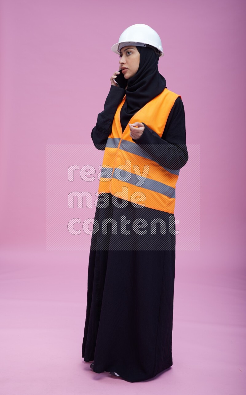 Saudi woman wearing Abaya with engineer vest and helmet standing talking on phone on pink background