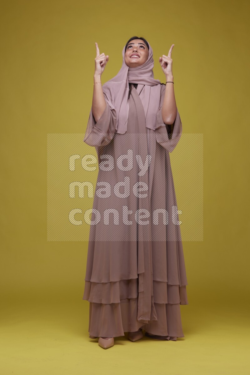 A woman Pointing on a Yellow Background wearing Brown Abaya with Hijab