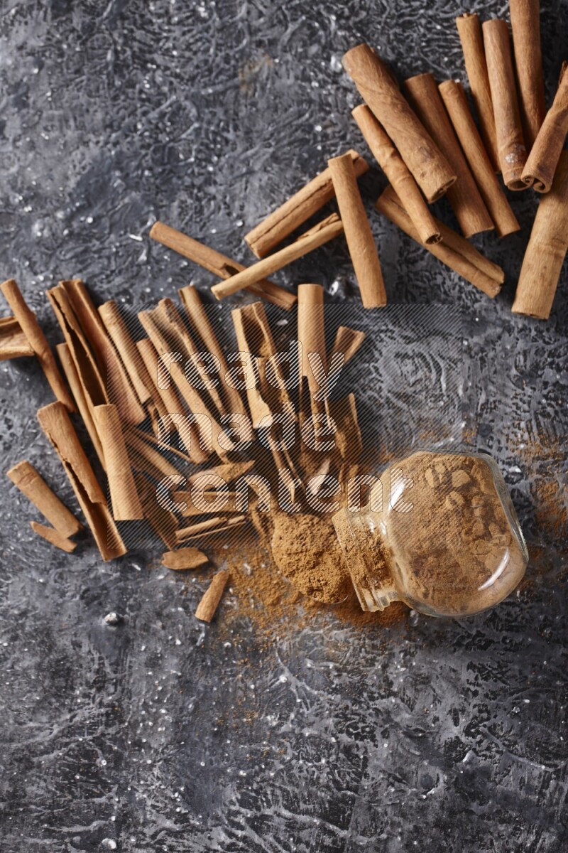 Herbal glass jar full cinnamon powder flipped and a metal spoon full of powder surrounded by cinnamon sticks on textured black background in different angles