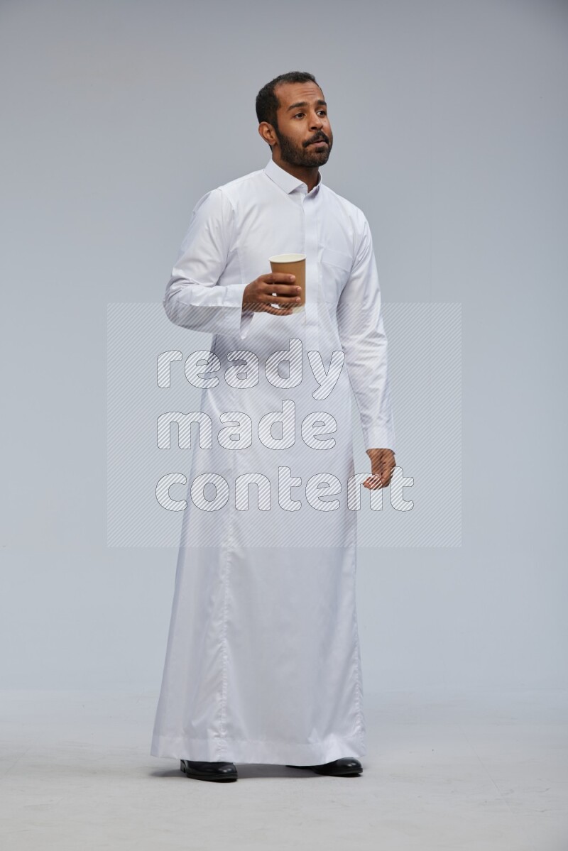 Saudi man wearing thob standing holding paper cup on gray background