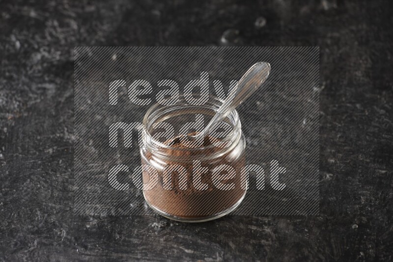 A glass jar full of cloves powder with a metal spoon on a textured black flooring