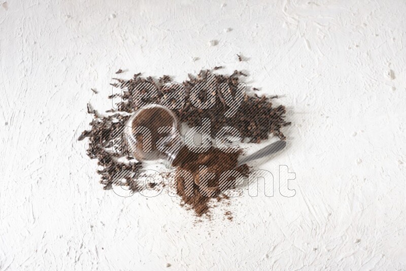 وعاء زجاجي مقلوب ممتلئ ببودرة القرنفل وتناثرت البودرة منه مع حبوب القرنفل المنتشرة على أرضية بيضاء