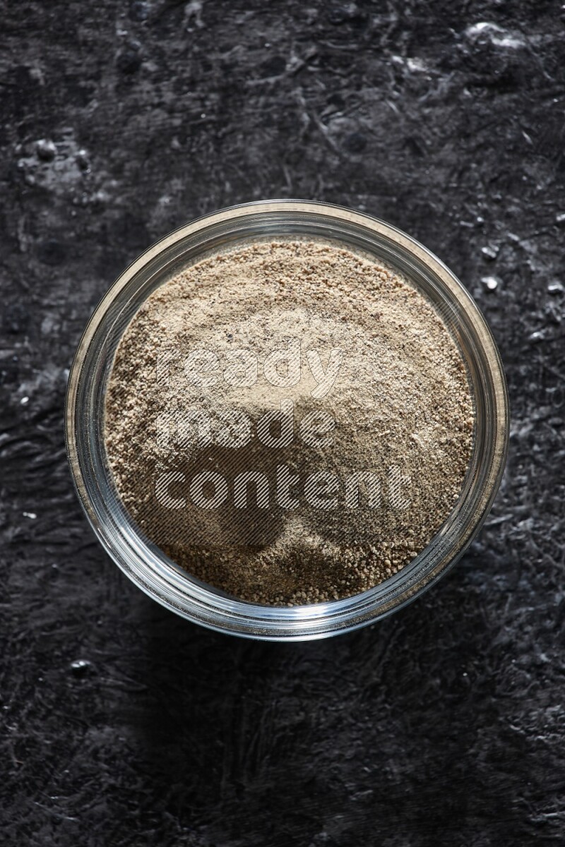 A glass bowl full of white pepper powder on textured black flooring