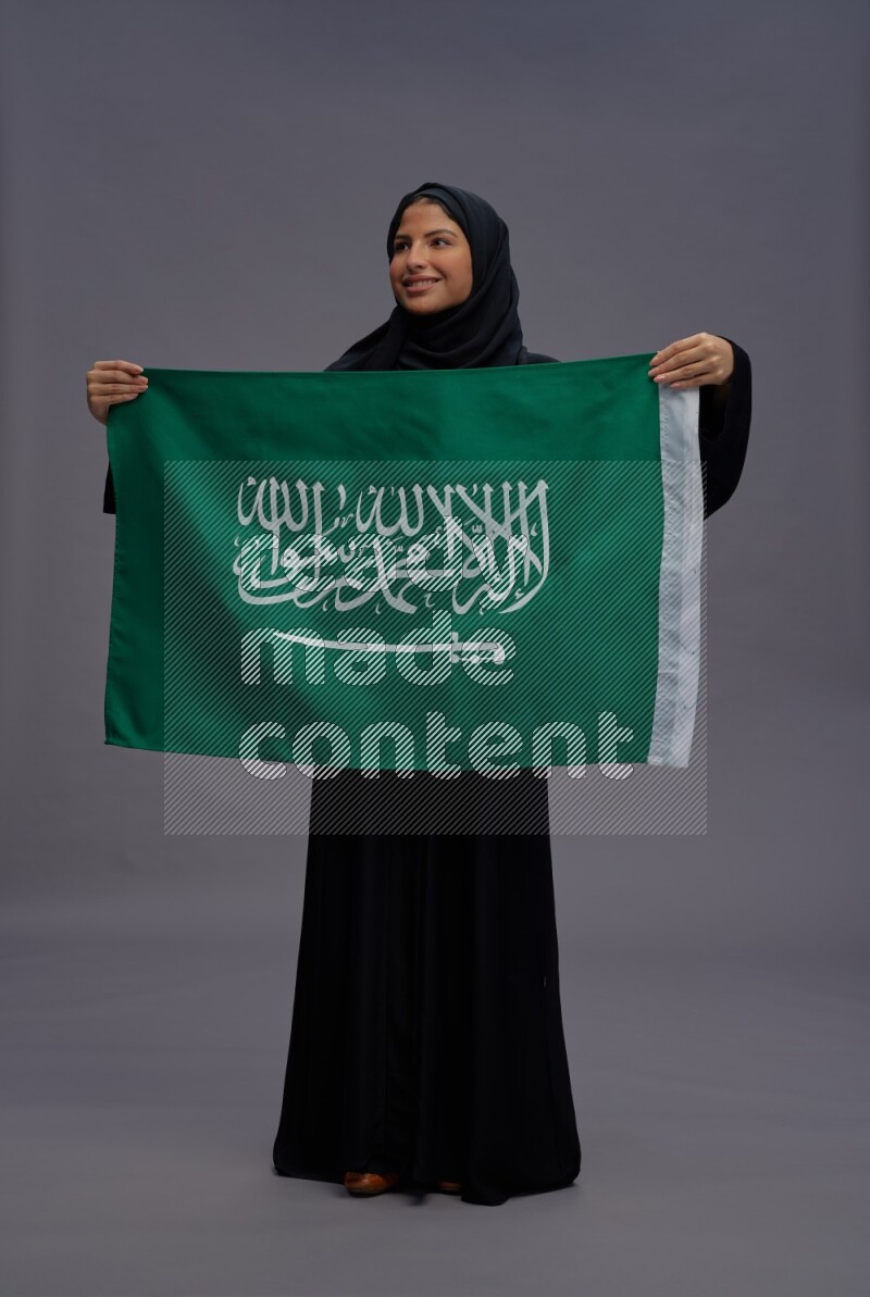Saudi woman wearing Abaya standing holding Saudi flag on gray background
