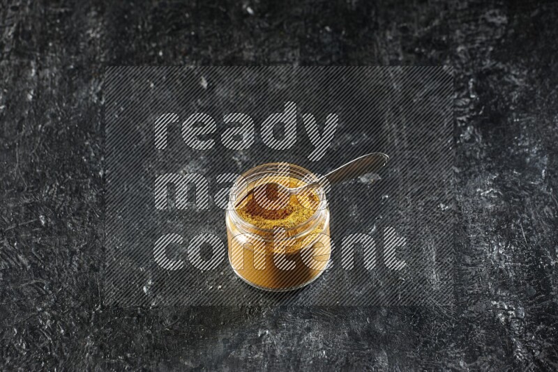 وعاء زجاجي وملعقة معدنية ممتلئان ببودرة الكركم على خلفية سوداء