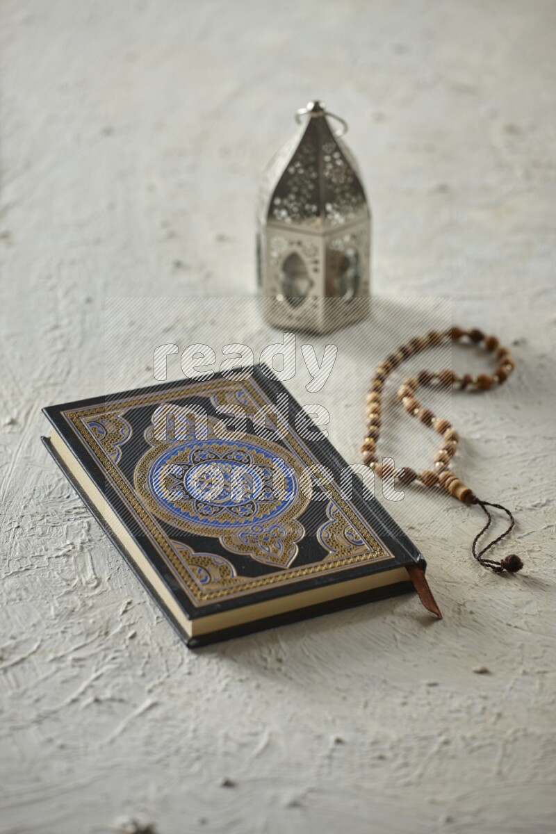 A silver lantern with different drinks, dates, nuts, prayer beads and quran on textured white background