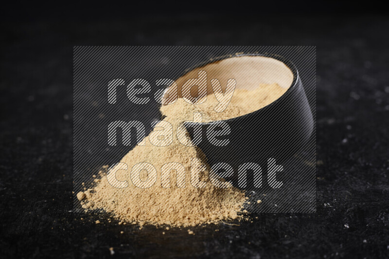 A black pottery bowl full of ground ginger powder with fallen powder from it on black background