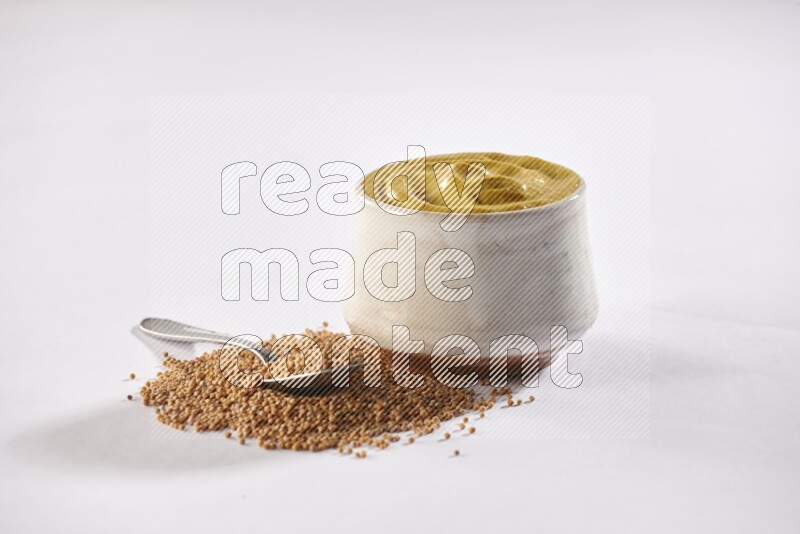 A beige pottery bowl full of mustard paste with mustard seeds underneath with a metal spoon on white flooring