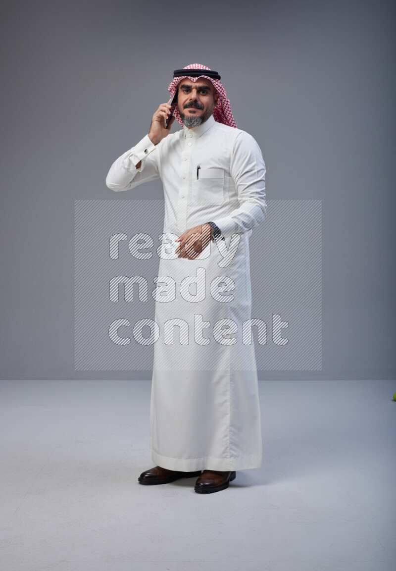 Saudi man Wearing Thob and red Shomag standing talking on phone on Gray background