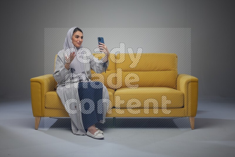 A Saudi woman wearing a light gray Abaya and head scarf sitting on a yellow sofa and making a video call eye level on a grey background