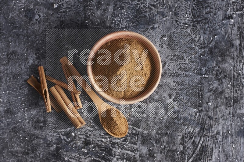 wooden bowl full of cinnamon powder and a wooden spoon full of it with cinnamon sticks on a textured black background