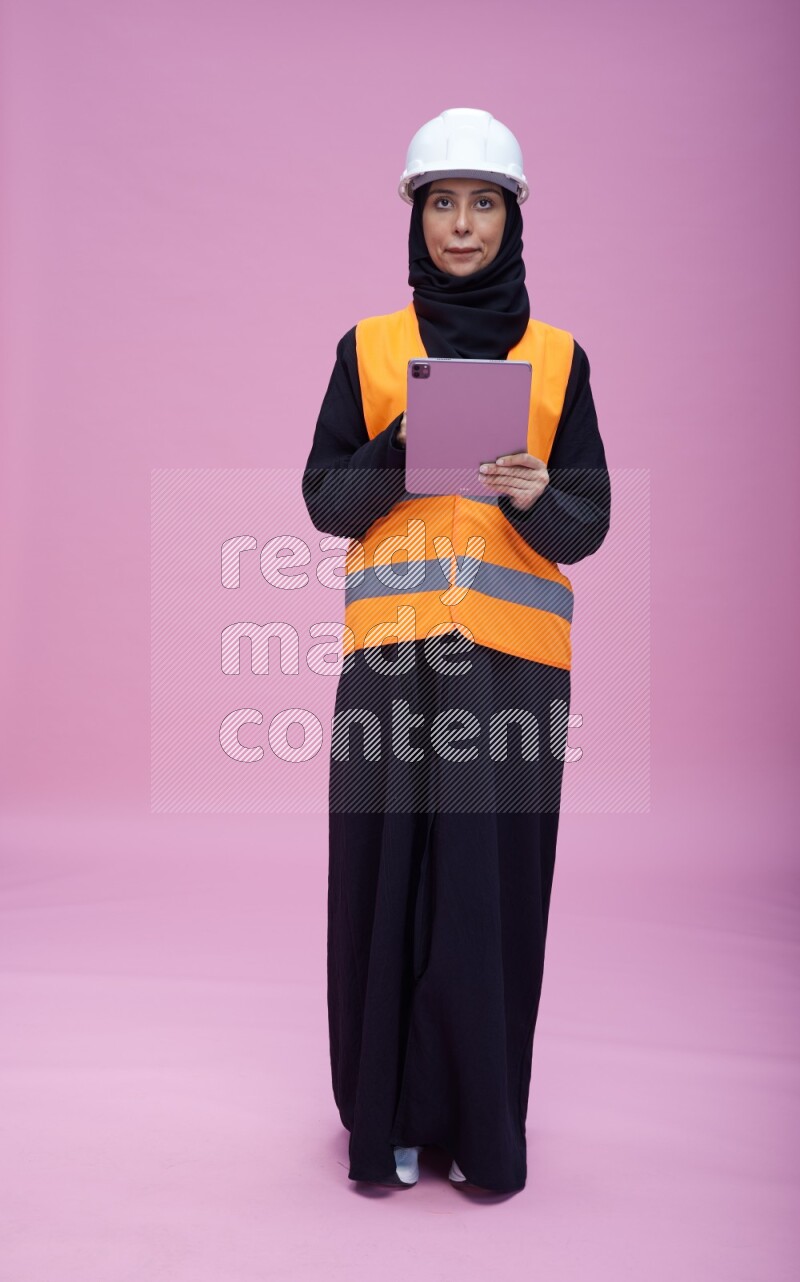 Saudi woman wearing Abaya with engineer vest and helmet standing working on tablet on pink background