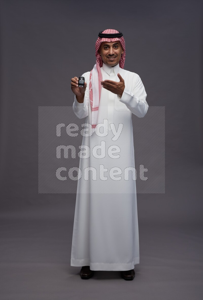 Saudi man wearing thob and shomag standing holding key car on gray background
