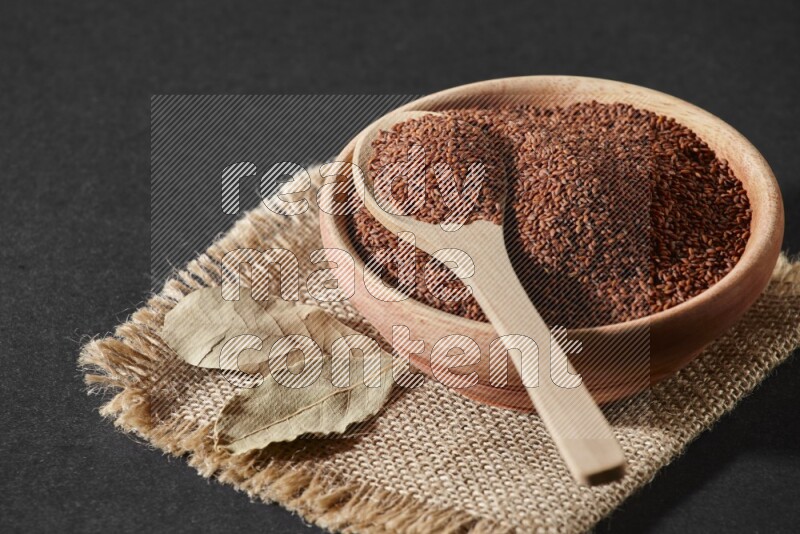 A wooden bowl full of garden cress seeds with wooden spoon full of the seeds on it on burlap fabric on a black flooring