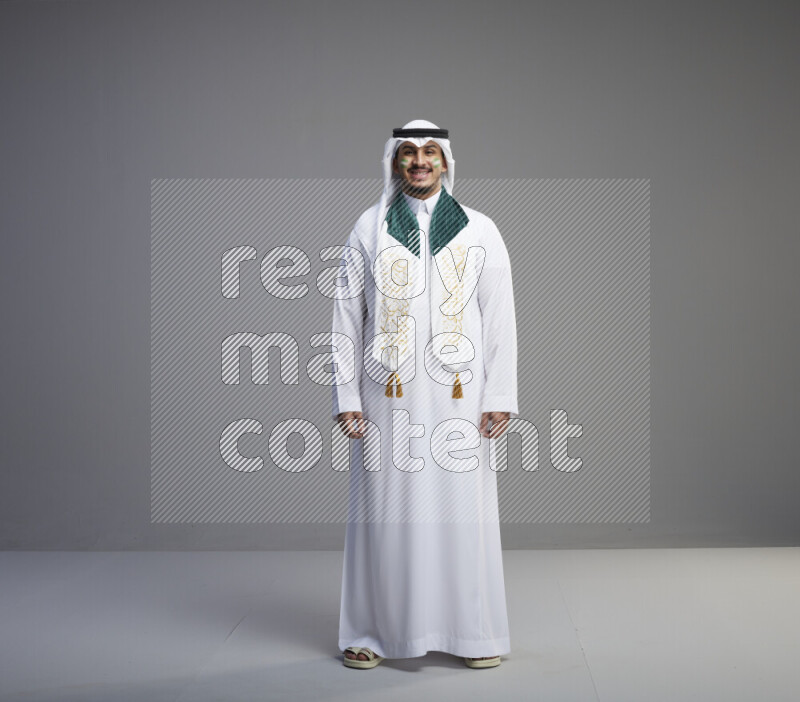 A Saudi man standing wearing thob and white shomag with face painting and Saudi flag scarf on gray background