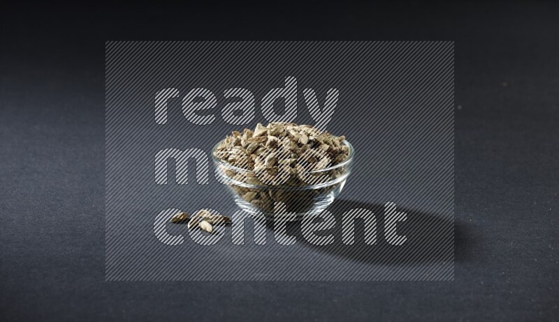A glass bowl full of cardamom and more seeds spreaded beneath the bowl on black flooring