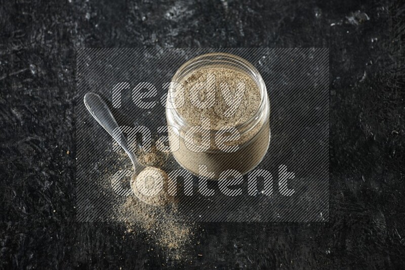 A glass jar and metal spoon full of cardamom powder on textured black flooring